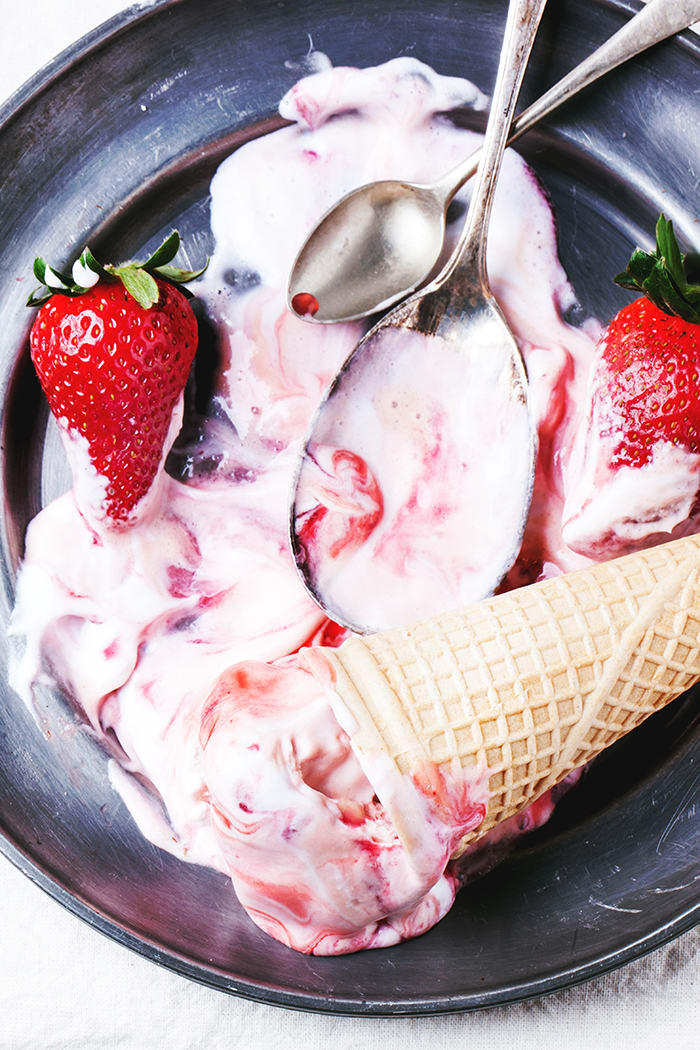 Wafer cones with strawberry ice cream with syrup, spoons and fresh strawberries served on vintage plate over white textile. Top view.